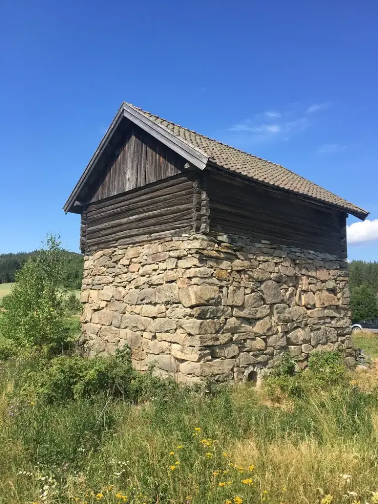 Bygning med høy mur av naturstein i åkerlandskap. På toppen av ovnen er det bygd en tømmerkasse med saltak. 
