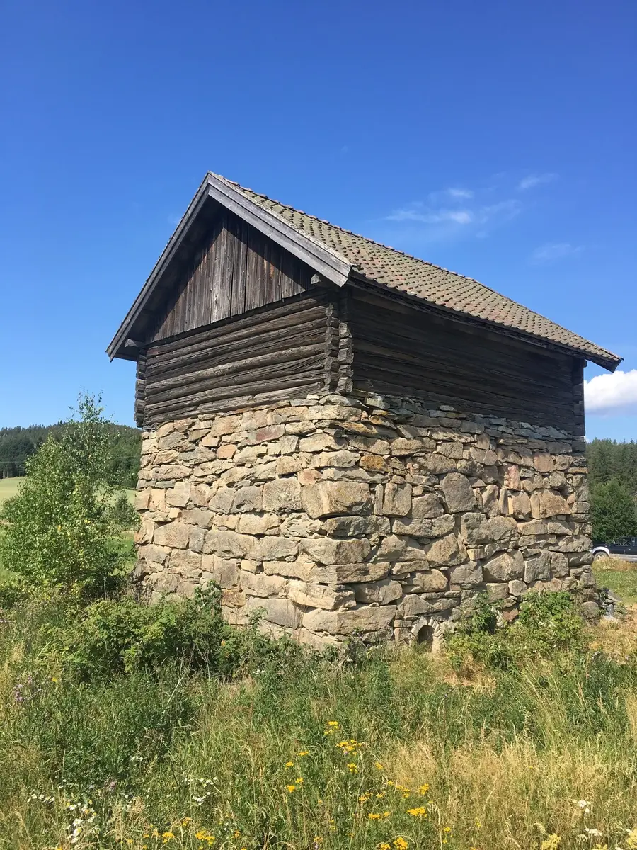 Bygning med høy mur av naturstein i åkerlandskap. På toppen av ovnen er det bygd en tømmerkasse med saltak. 