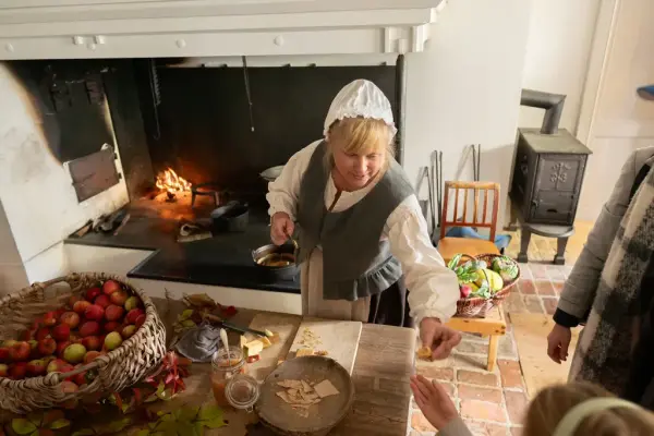 En kvinne deler ut smaksprøver i kjelleren på Eidsvollsbygningen. Det er fyr i grua og en kurv med epler på bordet. 