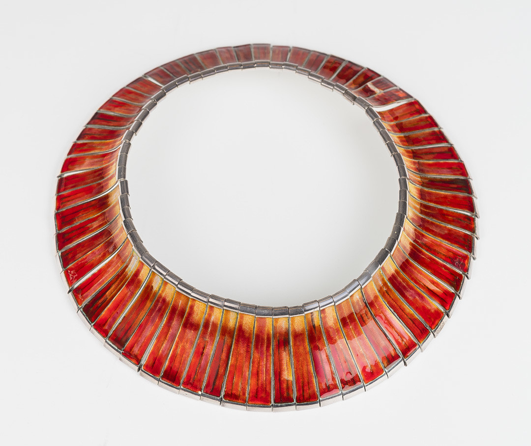 Halssmykke i sølv og speilemalje. Det er bygget opp av rektangulære ledd i sølv, hvis overflate er håndfreset og dekket med rødlig og gulfarget emalje.