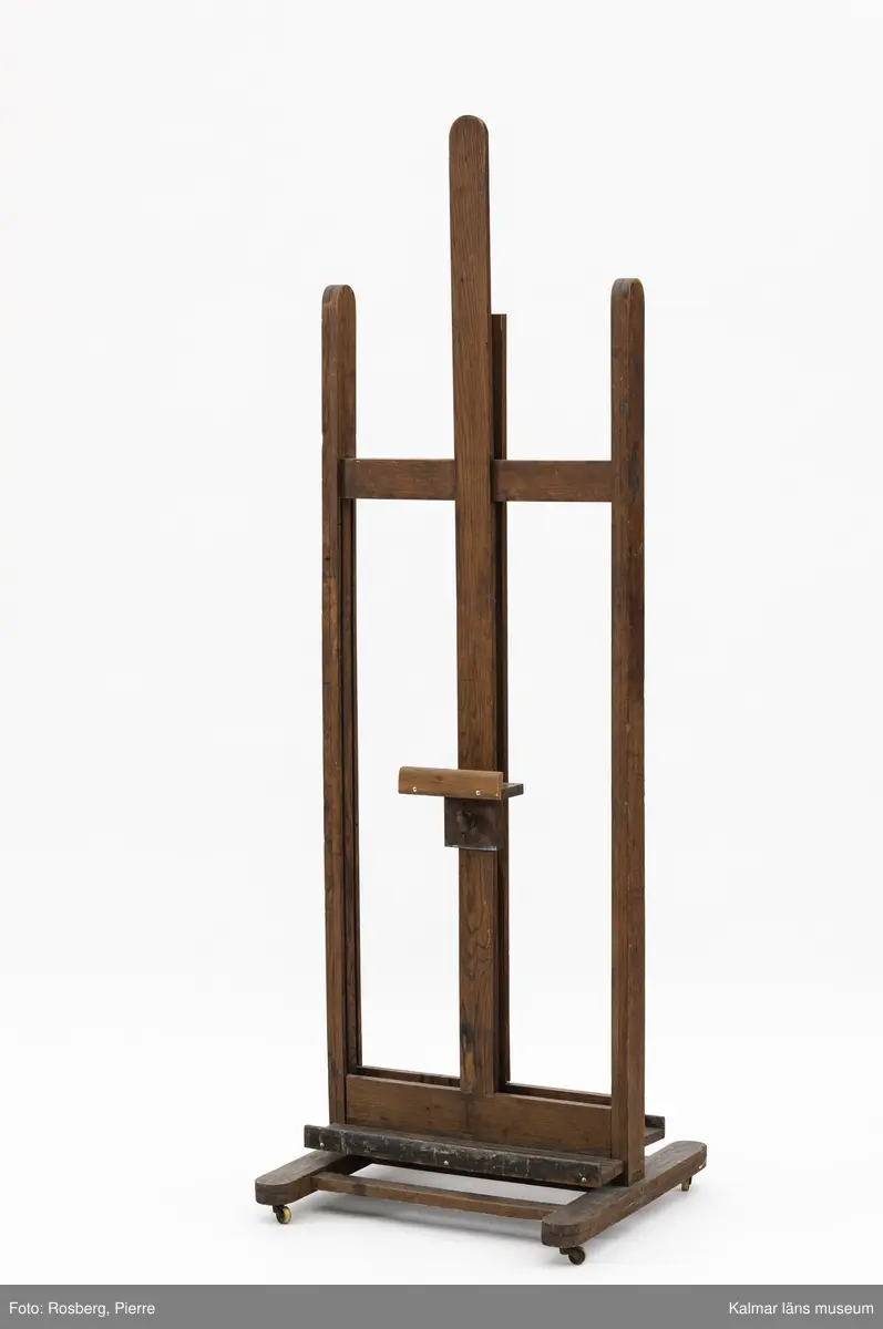 KLM 31949. Staffli. Tillverkat av ek. Staffliet kan användas från båda sidor. Staffliet består av tre parallella stolpar. På den mittersta stolpen löpes tavelstödet. Nedtill på båda sidorna finns en hylla för penslar, färg m.m. Staffliet går på fyra små mässingshjul.