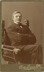 En man i mörk kostym med väst och stärkkrage med fluga, sitt