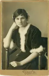 En ung kvinna i tankfull pose, klädd i mörk kjol och mörk sammetsblus kantad med svandun. Hon sitter i en s.k. munkstol. 
Text: "Greta" nedtill, + oläslig, präglad fotografsignatur. 
Knäbild, halvprofil. Ateljéfoto.