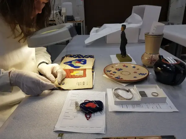 Fotografi av et bord. Til venstre for bordet står ei kvinne lent over ei bok som ligger på bordet, hun kikker gjennom et slags forstørrelsesglass med lys. På bordet ligger det ulike gjenstander.