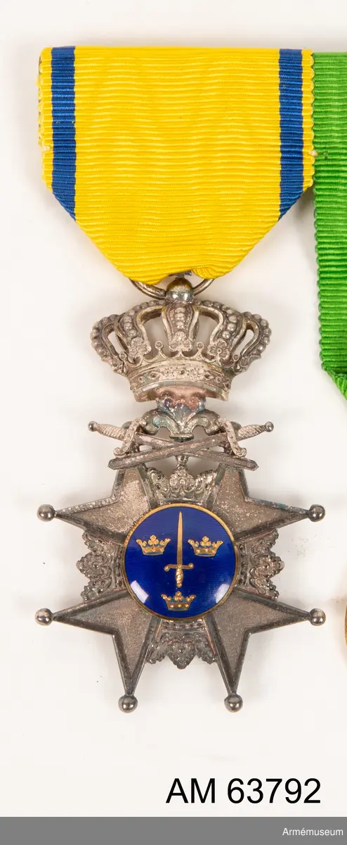 Grupp M II.
Svärdsmannatecken med band tilldelat styckjunkaren Georg Wilhelm Wessman.  Ett snedställt johannitkors av silver. Åtta uddar, var och en slutande i en kula. Mellan korsarmarna öppna kronor. På åtsidan har tecknet en blå, emaljerad glob i korstets mitt med ett stolpvis ställt svärd och tre öppna kronor ställda två och en. Mellan korsets övre vänstra udd och övre högra störtande svärd. Frånsidans mittglob är av silver med ett stolpvis ställt svärd omlagt med två kvistar ovanför "PRO PATRIA". Tecknet hänger i en kunglig krona. Medaljband av ett 35 mm brett sidenripsband - gult med mörkblå kantrand. 