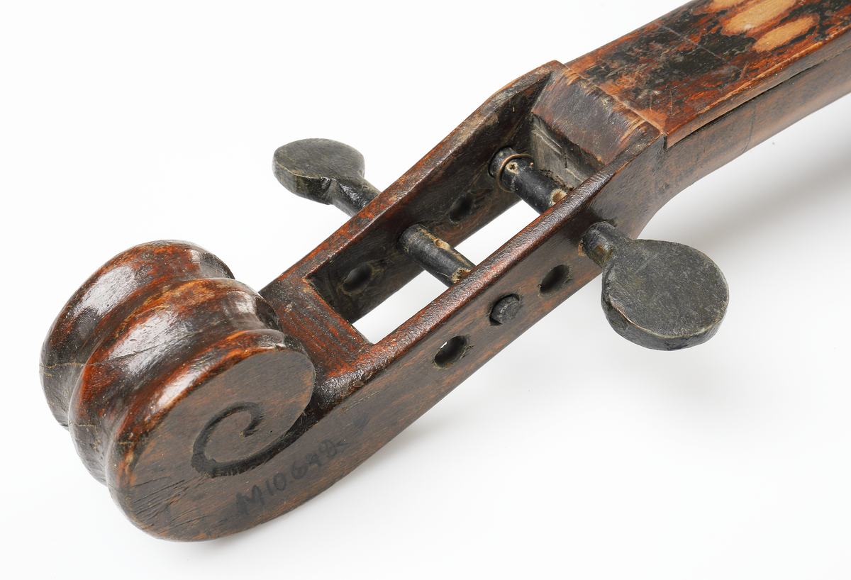 Fiol av allmogetillverkning, helt gjord av trä. Fiolen har två svartmålade stämskruvar. Ljudhålen på locket är f-formade. Strängfäste, stall, strängar och två stämskruvar saknas. Tillhörande stråke.