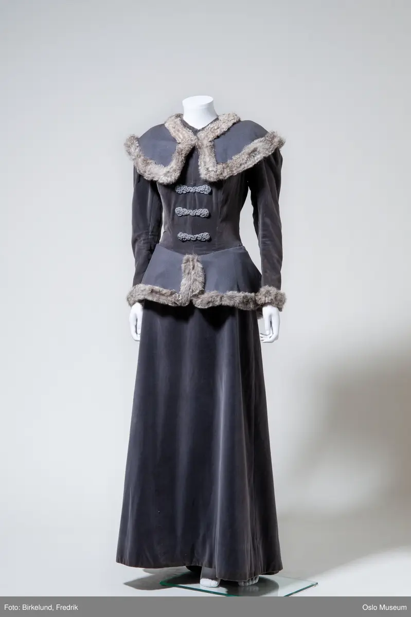 Lang grå draktkjole i ull med lage ermer og kappedekor på skuldre og hofter. Pelsdekor langs kantene.
Underskjørt