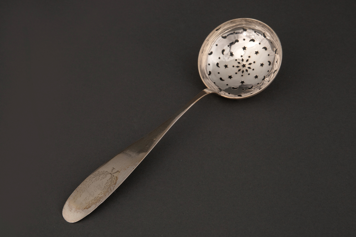 Strøskje i sølv med sirkulært, øseformet blad med buet bunn. Bunnen er gjennombrutt med runde, diamant-, stjerne- og dråpeformede hull. Skaftet tiltar i bredde mot en avrundet avslutning. Ytterst på skaftet er det gravert enkel bladdekor.