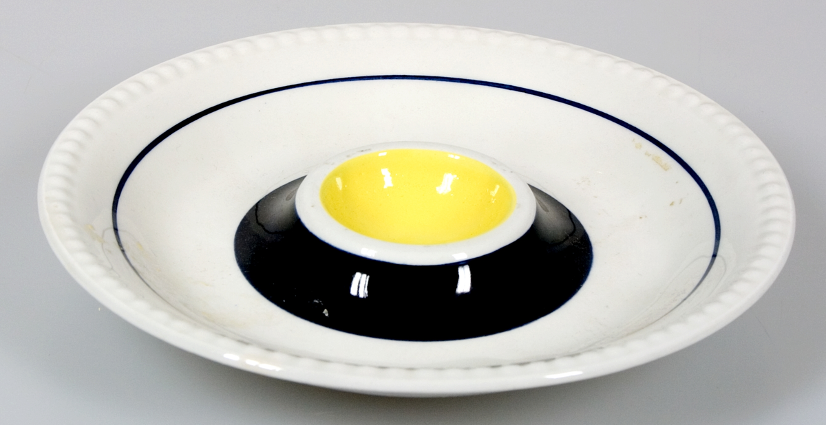 Två äggkoppar av vitglaserat porslin utformade som assietter med "kopp" för ägg i mitten. Dekor i gult och mörkblått. Stämplade: Upsala-Ekeby, UE, SWEDEN, GEFLE.
