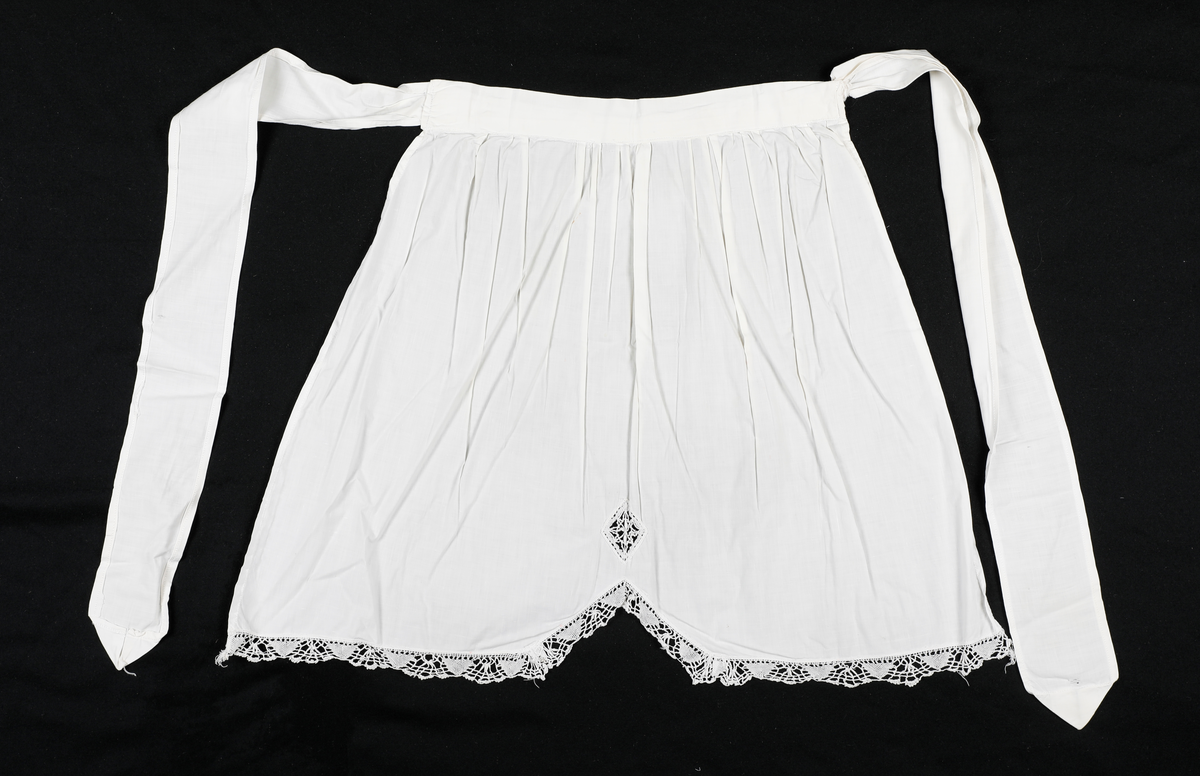 Forkle med påsydd blondekant nederst. Nedkanten på forkleet går opp i en spiss på midten.