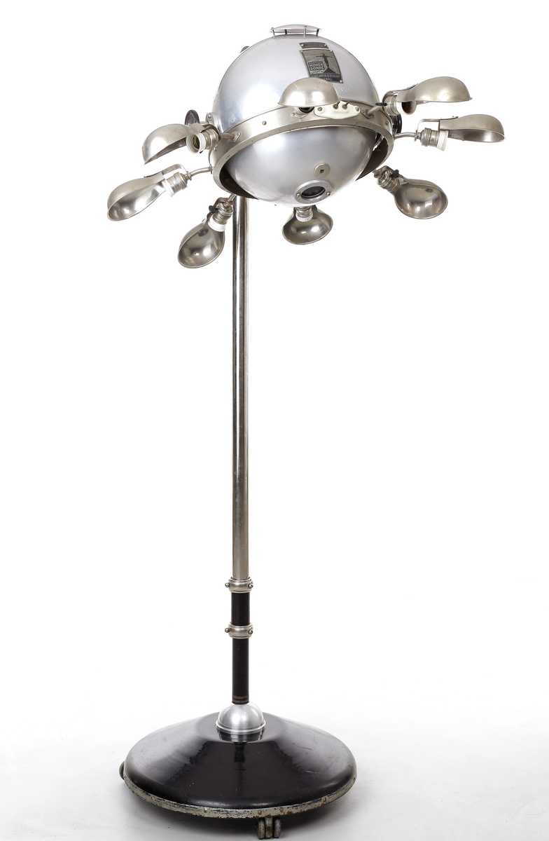 Kvartslampa som består av en förnicklad pelare med en höj- och sänkbar lampa. Lampan har ett klot vars innehåll  är en kvartslampa med kvicksilver som bildar UV-ljus. Huven har en öppningsbar del som kan öppnas med en låsbar ratt på ena sidan så själva lamphuset kan kommas åt. Denna öppningsbara del har en vridbar skiva med hål i olika storlek, 15-ca. 45mm, detta så att man kan ha olika koncentration på ljusstrålen. På andra sidan finns en motsvarande ratt som man kan justera själva kvartslampan. Runt om klotet sitter åtta stycken skärmförsedda lamphållare där varannan lamphållaren har en vridströmbrytare så att varanna lampa kan släckas. Dessa lamphållare har lampfästen i vitt porslin, och vridströmbrytarna är troligen i bakelit. Det sitter även två stycken elkontakter i porslin på klotets utsida. Foten på lampan består av en cirkelformad svartlackerad välvd platta med tre stycken vridbara dubbelhjul. På huvens utsidan sitter även två tillverkarskyltar fastnitade som har texten "QUARZLAMPEN-GESELLSCHAFT m.b.H. HANAU No. 38673, 7,8/3,8 Amp. 110/220 Volt." och "KÜNSTLICHE HÖHENSONNE QUARZLAMPEN-GESELLSCHAFT HANAU".