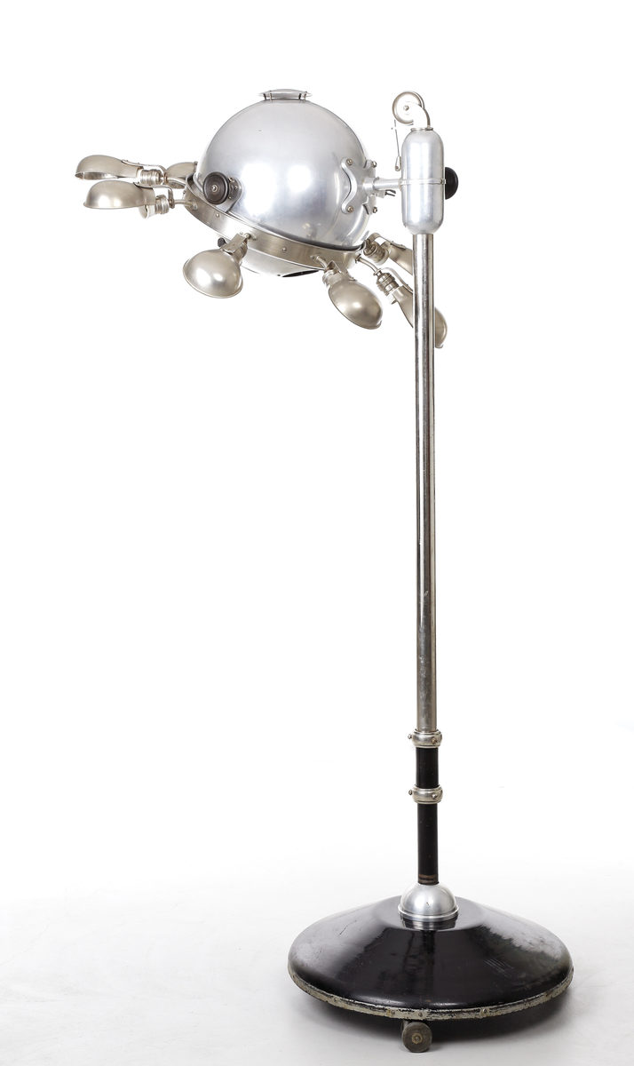 Kvartslampa som består av en förnicklad pelare med en höj- och sänkbar lampa. Lampan har ett klot vars innehåll  är en kvartslampa med kvicksilver som bildar UV-ljus. Huven har en öppningsbar del som kan öppnas med en låsbar ratt på ena sidan så själva lamphuset kan kommas åt. Denna öppningsbara del har en vridbar skiva med hål i olika storlek, 15-ca. 45mm, detta så att man kan ha olika koncentration på ljusstrålen. På andra sidan finns en motsvarande ratt som man kan justera själva kvartslampan. Runt om klotet sitter åtta stycken skärmförsedda lamphållare där varannan lamphållaren har en vridströmbrytare så att varanna lampa kan släckas. Dessa lamphållare har lampfästen i vitt porslin, och vridströmbrytarna är troligen i bakelit. Det sitter även två stycken elkontakter i porslin på klotets utsida. Foten på lampan består av en cirkelformad svartlackerad välvd platta med tre stycken vridbara dubbelhjul. På huvens utsidan sitter även två tillverkarskyltar fastnitade som har texten "QUARZLAMPEN-GESELLSCHAFT m.b.H. HANAU No. 38673, 7,8/3,8 Amp. 110/220 Volt." och "KÜNSTLICHE HÖHENSONNE QUARZLAMPEN-GESELLSCHAFT HANAU".