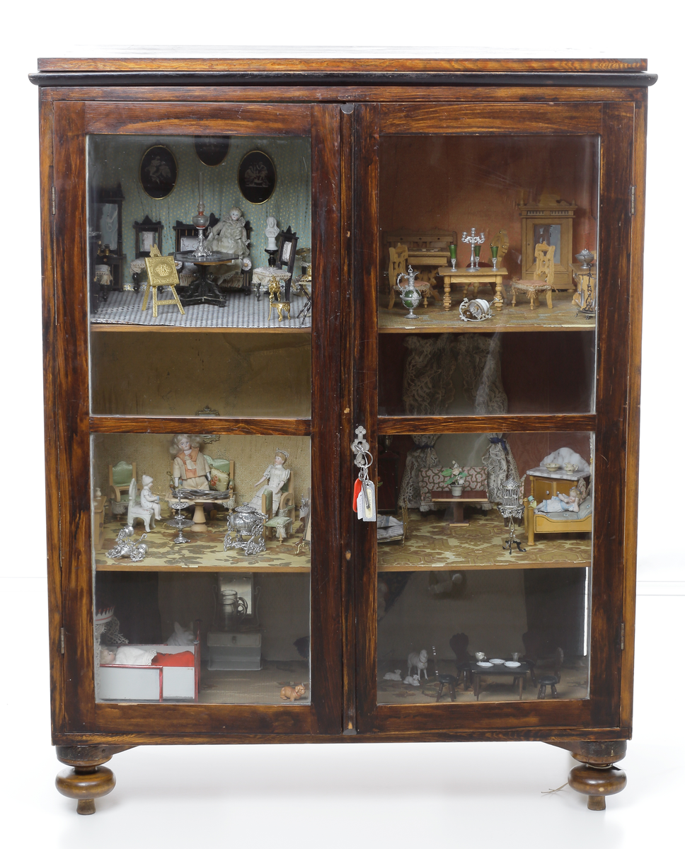 Dockskåp med tillhörande inredning. Brunbetsat träskåp försett med glasförsedda pardörrar. Skåpet har tre våningsplan med fem möblerade rum. Inredning från 1890-talet. Skåpet står på fyra svarvade fötter.