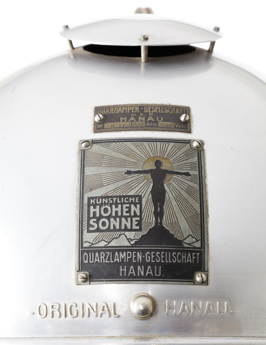 Kvartslampa som består av en förnicklad pelare med en höj- och sänkbar lampa. Lampan har ett klot vars innehåll  är en kvartslampa med kvicksilver som bildar UV-ljus. Huven har en öppningsbar del som kan öppnas med en låsbar ratt på ena sidan så själva lamphuset kan kommas åt. Denna öppningsbara del har en vridbar skiva med hål i olika storlek, 15-ca. 45mm, detta så att man kan ha olika koncentration på ljusstrålen. På andra sidan finns en motsvarande ratt som man kan justera själva kvartslampan. Runt om klotet sitter åtta stycken skärmförsedda lamphållare där varannan lamphållaren har en vridströmbrytare så att varanna lampa kan släckas. Dessa lamphållare har lampfästen i vitt porslin, och vridströmbrytarna är troligen i bakelit. Det sitter även två stycken elkontakter i porslin på klotets utsida. Foten på lampan består av en cirkelformad svartlackerad välvd platta med tre stycken vridbara dubbelhjul. På huvens utsidan sitter även två tillverkarskyltar fastnitade som har texten "QUARZLAMPEN-GESELLSCHAFT m.b.H. HANAU No. 38673, 7,8/3,8 Amp. 110/220 Volt." och "KÜNSTLICHE HÖHENSONNE QUARZLAMPEN-GESELLSCHAFT HANAU".