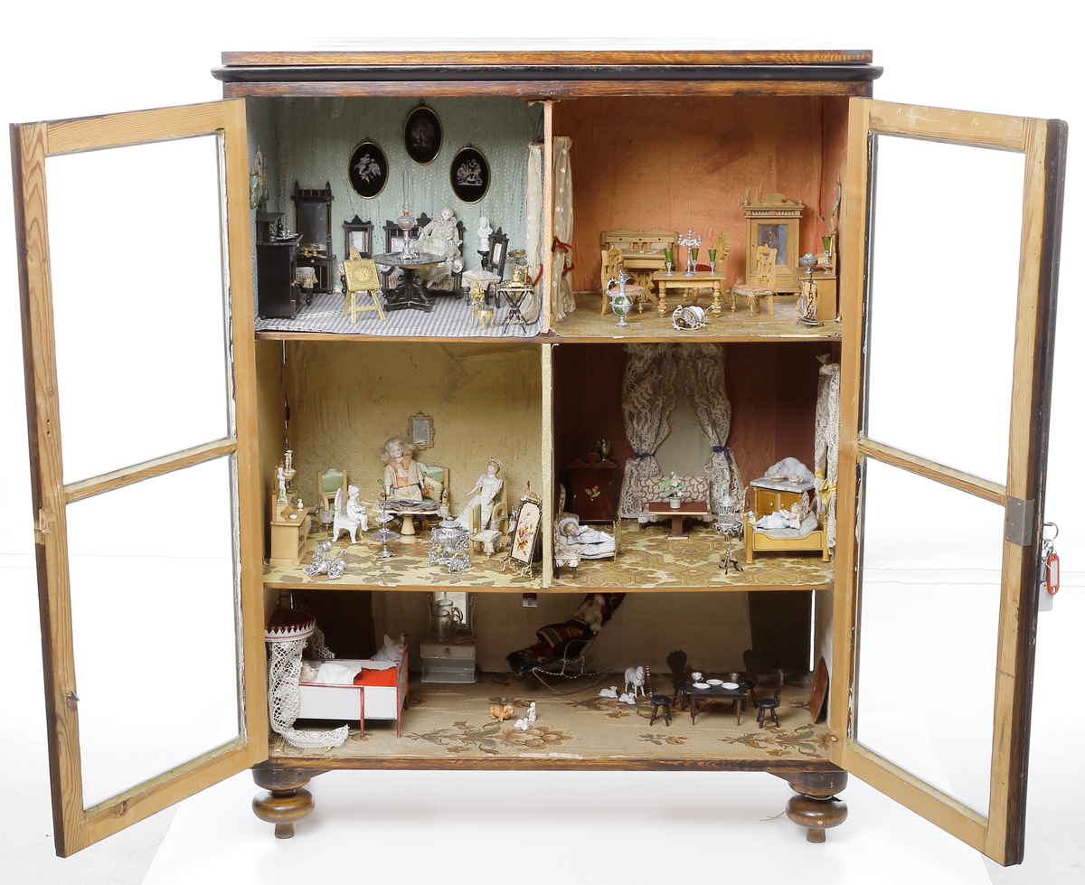 Dockskåp med tillhörande inredning. Brunbetsat träskåp försett med glasförsedda pardörrar. Skåpet har tre våningsplan med fem möblerade rum. Inredning från 1890-talet. Skåpet står på fyra svarvade fötter.