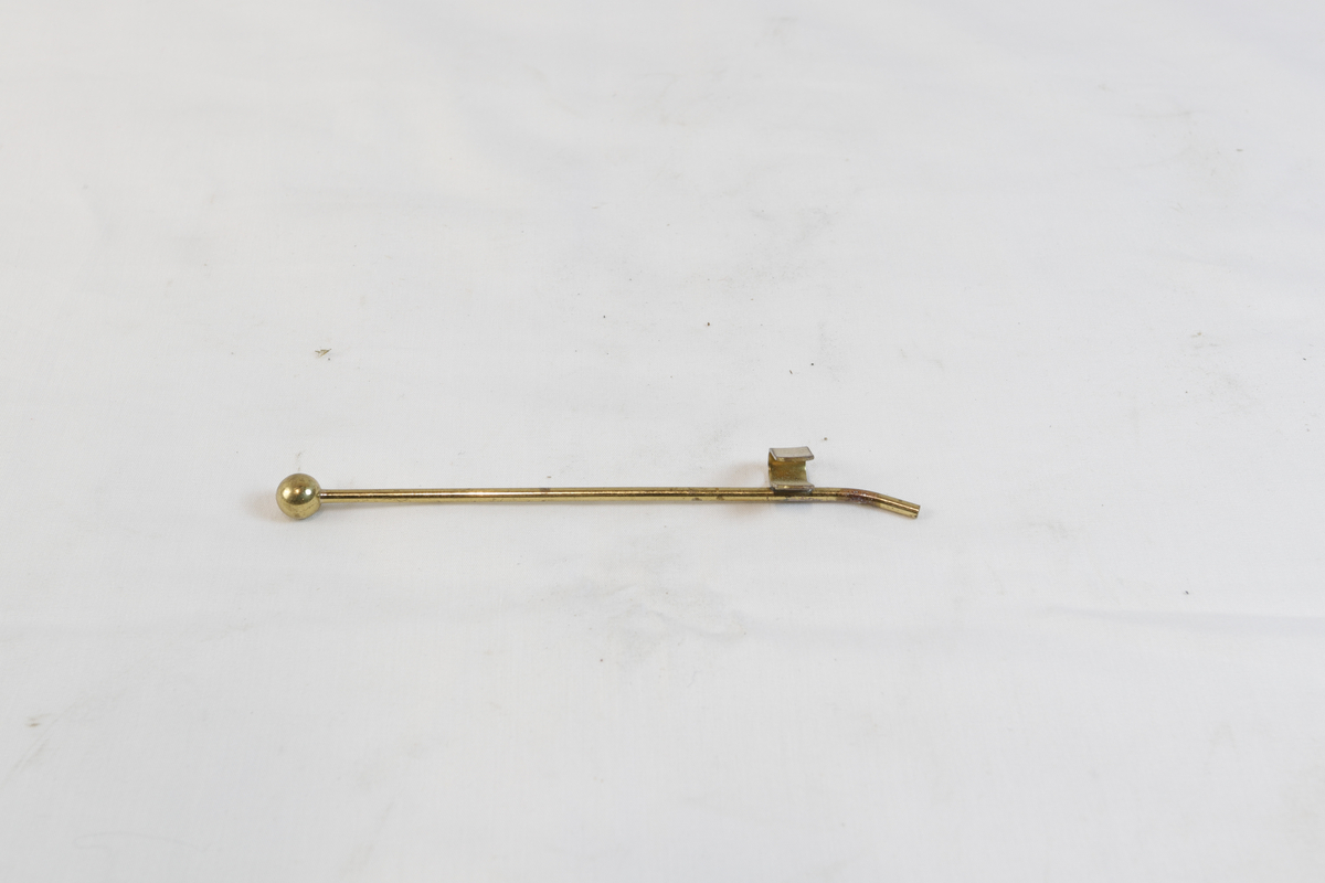 Lang gjenstand i gyllent metall. Den ene delen har en kule i enden og på den andre siden er det en hemp.