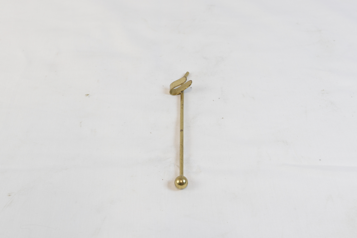 Lang gjenstand i gyllent metall. Den ene delen har en kule i enden og på den andre siden er det en hemp.