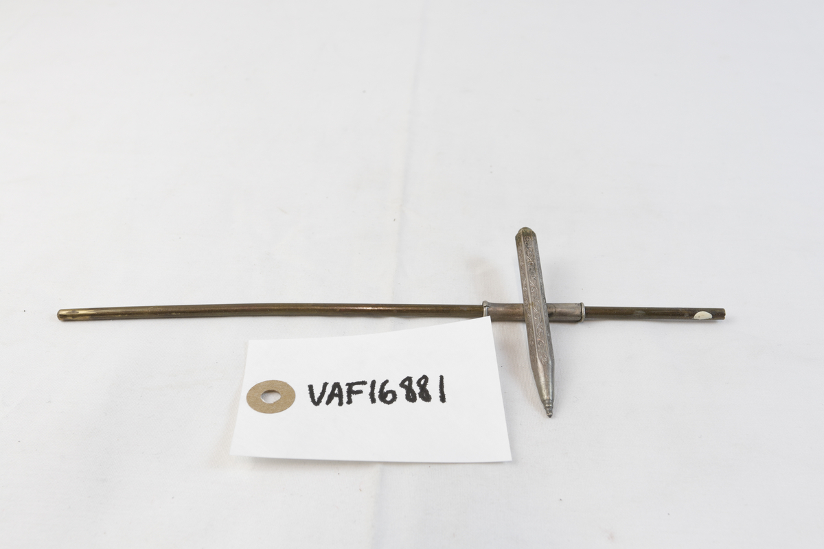 Blyant-formet gjenstand i sølvplett med inngravert mønster. Den er festet til en lengre stang i metall. 