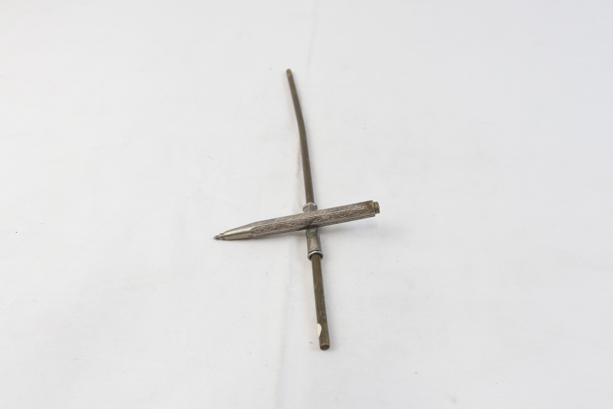 Blyant-formet gjenstand i sølvplett med inngravert mønster. Den er festet til en lengre stang i metall. 