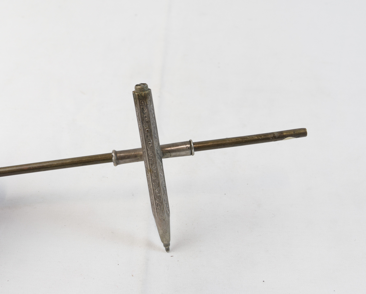 Blyant-formet gjenstand i sølvplett med inngravert mønster. Den er festet til en lengre stang i metall. 