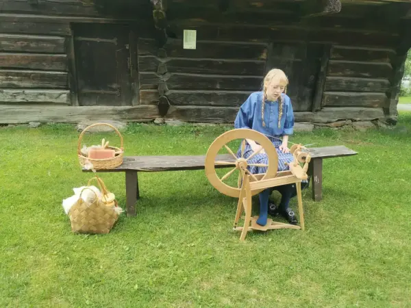 Jente som spinner ull foran eit gamalt hus