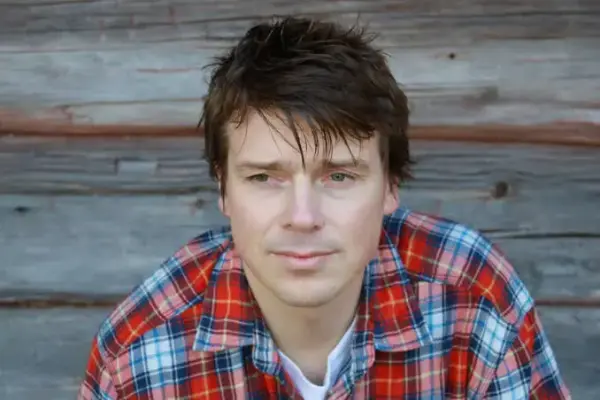 Bilde av Øystein Morten. Portrettbilde av mann med mørkt hår og rutete skjorte foran en tømmervegg