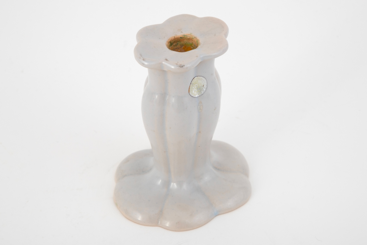 Lysestake i keramikk. Lysestaken har en oval base som går over i et skaft som smalner noe inn mot midten. Øverst er kanten fortykket. Lysestakten er dekorert med langsgående, jevnt fordelte dype rifler, noe som gir den et organisk uttrykk. Glasert i hvitt med blåskjær.