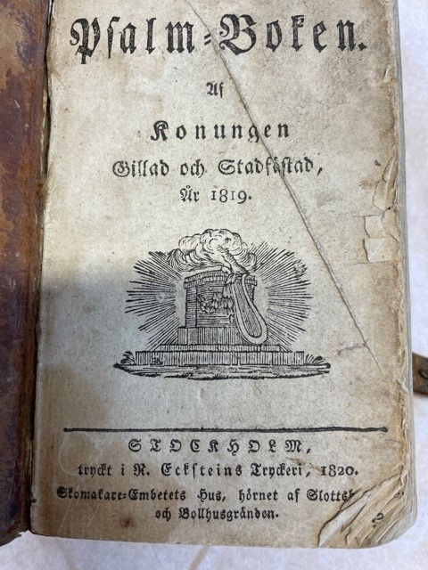 Psalmbok, tryckt i Stockholm, helfranskt band, pärm skadad men ursprungligen klädd med papper. Försättsblad med titel 
"Den Swenska Psalm-Boken. af Konungen Gillad och Stadfästad, år 1819. STOCKHOLM, tryckt i R. Ecksteins Tryckeri, 1820. Skomakare-Embetets Hus, hörnet af Slottsbacken och Bollhusgränden".  Försättsblad dekorerad med tryck föreställande postamen, och villande lyra samt rökformation. På baksidan del av spänne av läder med fäste i mässing

