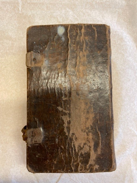 Psalmbok, tryckt i Stockholm, helfranskt band, pärm skadad men ursprungligen klädd med papper. Försättsblad med titel 
"Den Swenska Psalm-Boken. af Konungen Gillad och Stadfästad, år 1819. STOCKHOLM, tryckt i R. Ecksteins Tryckeri, 1820. Skomakare-Embetets Hus, hörnet af Slottsbacken och Bollhusgränden".  Försättsblad dekorerad med tryck föreställande postamen, och villande lyra samt rökformation. På baksidan del av spänne av läder med fäste i mässing

