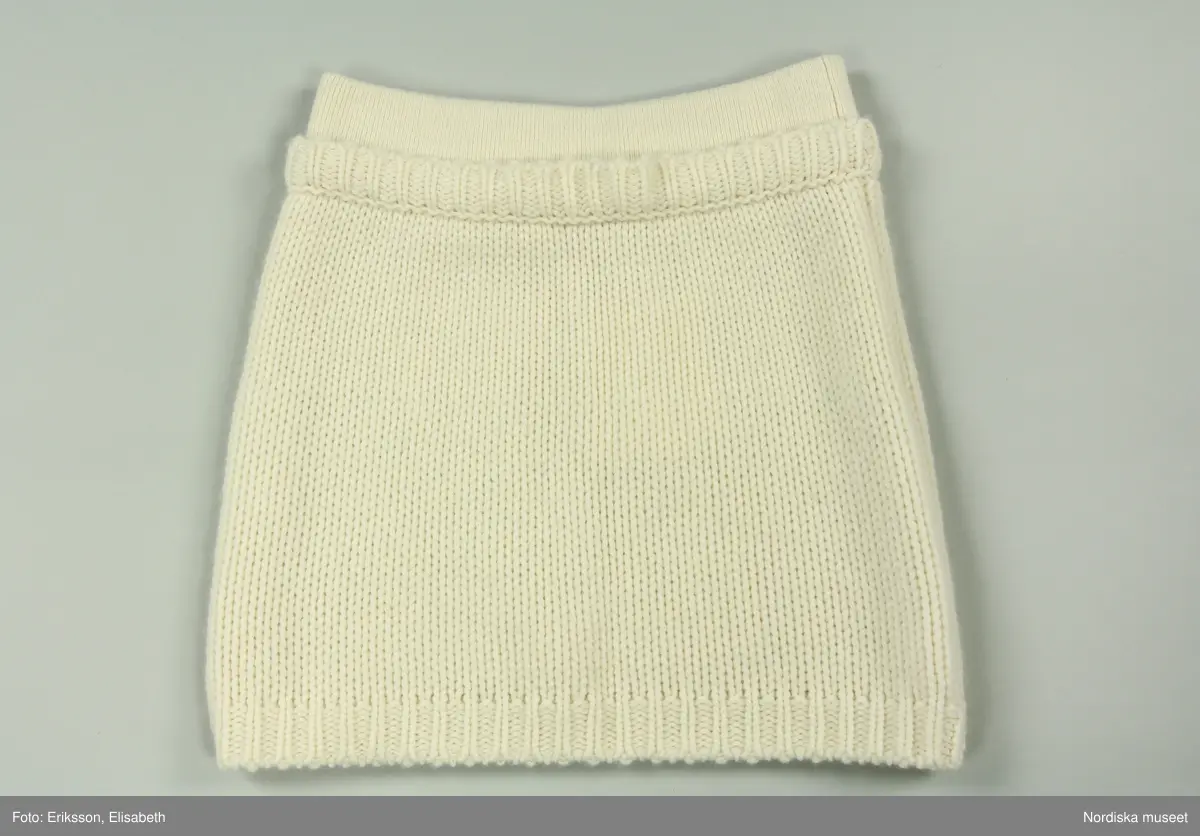 Kjol, stickad av vitt kashmirgarn. Kort modell. Upptill resårstickad linning, strax därunder smal, stickad, horisontal remsa, vikt uppåt. Nedtill dekorstickning.  
På insidan av linningen (bak) vit tygetikett med text: LISA YANG". Vid denna etikett fäst med vitt snöre två pappersetiketter.
Fastknutet med vitt snöre i etiketten bak två pappersetiketter. Den ena med text "LISA YANG. Den andra med kinesiskt tecken (använt som firmans symbol) samt text: 
"Style. Lovi / Colour: Cream / Size: 1 / Style no.: 2023326".
Vid linningen också fäst tre vita tygetiketter med information om tillverkning, material, tvätt mm. samt liten etikett med storleksangivelse (1).
/Leif Wallin 2025-03-05