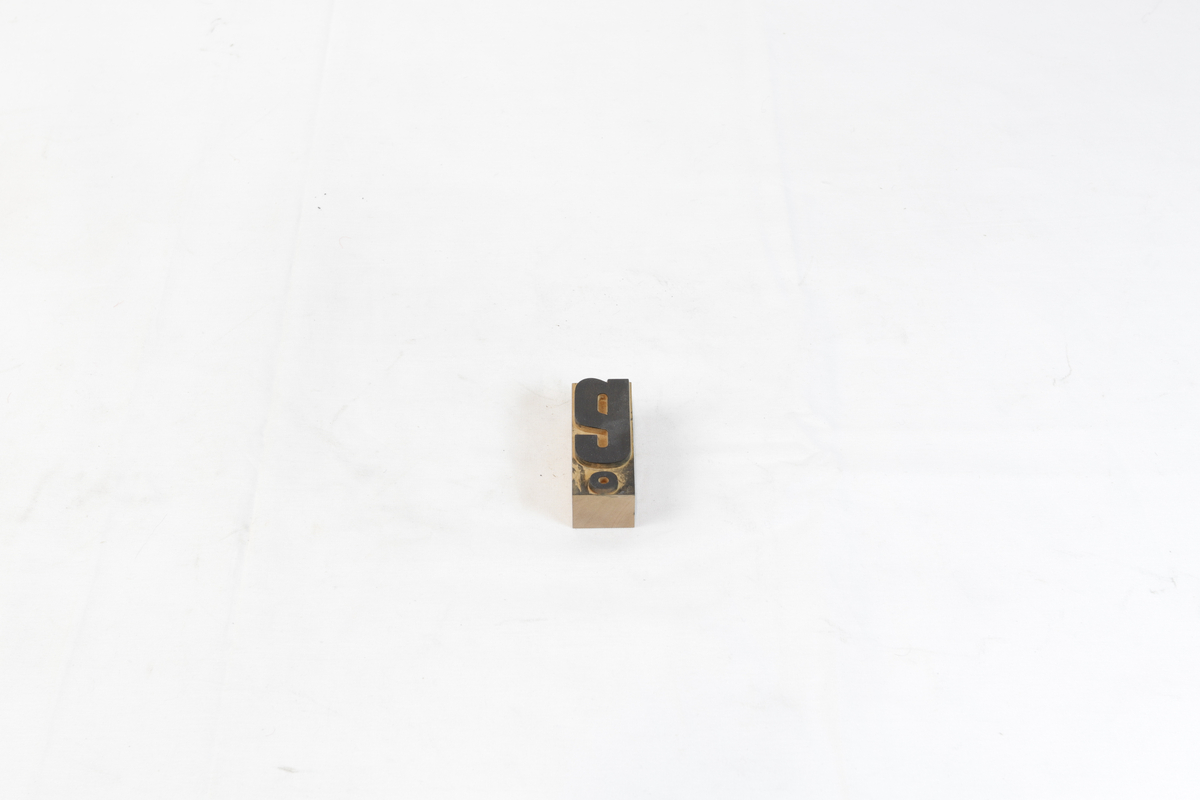 Stempel i tre. Stempelet har en rektangulær form med motiv av en litenbokstav. Bokstav: å. Det er svart trykksverte på stempelet.