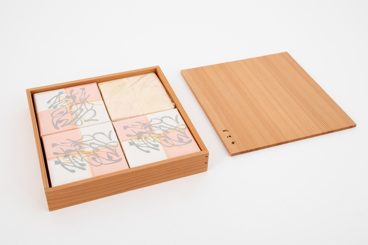 Lav kvadratisk treeske med flatt, uprofilert lokk. I esken ligger fire pakninger med fire kvadratiske konfektstykker i hver, plassert på små plater i japanseder. Innpakningspapiret er hvitt og rosa. Gullsnor rundt pakningene. Konfekten er laget av presset melis. Treesken ligger i en eske med lokk i kartong.