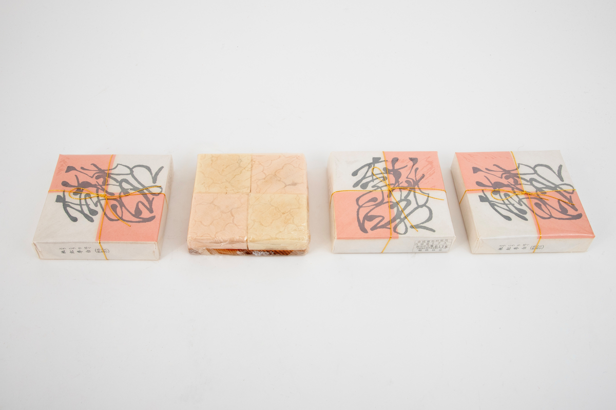 Lav kvadratisk treeske med flatt, uprofilert lokk. I esken ligger fire pakninger med fire kvadratiske konfektstykker i hver, plassert på små plater i japanseder. Innpakningspapiret er hvitt og rosa. Gullsnor rundt pakningene. Konfekten er laget av presset melis. Treesken ligger i en eske med lokk i kartong.