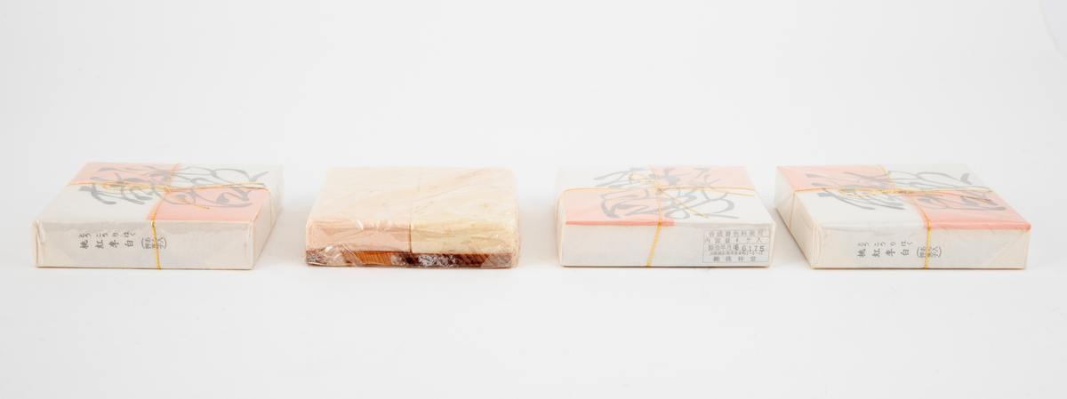 Lav kvadratisk treeske med flatt, uprofilert lokk. I esken ligger fire pakninger med fire kvadratiske konfektstykker i hver, plassert på små plater i japanseder. Innpakningspapiret er hvitt og rosa. Gullsnor rundt pakningene. Konfekten er laget av presset melis. Treesken ligger i en eske med lokk i kartong.