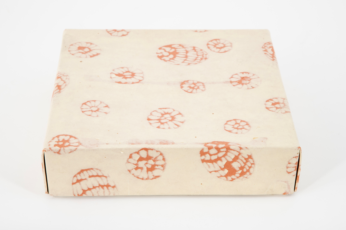 Lav kvadratisk treeske med flatt, uprofilert lokk. I esken ligger fire pakninger med fire kvadratiske konfektstykker i hver, plassert på små plater i japanseder. Innpakningspapiret er hvitt og rosa. Gullsnor rundt pakningene. Konfekten er laget av presset melis. Treesken ligger i en eske med lokk i kartong.