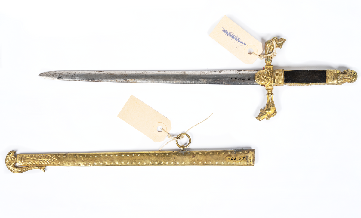 Stickert, oslipad klinga av plattoval form, handtag av ebenholtz beslaget med förgylld ciselerad mässing med manshuvud. Parerstång med två djurhuvuden. Balja av förgylld mönstrad mässing med lilla riksvapnet. En ring saknas.