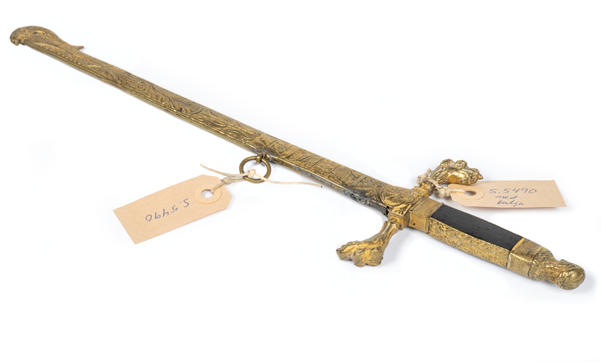 Stickert, oslipad klinga av plattoval form, handtag av ebenholtz beslaget med förgylld ciselerad mässing med manshuvud. Parerstång med två djurhuvuden. Balja av förgylld mönstrad mässing med lilla riksvapnet. En ring saknas.