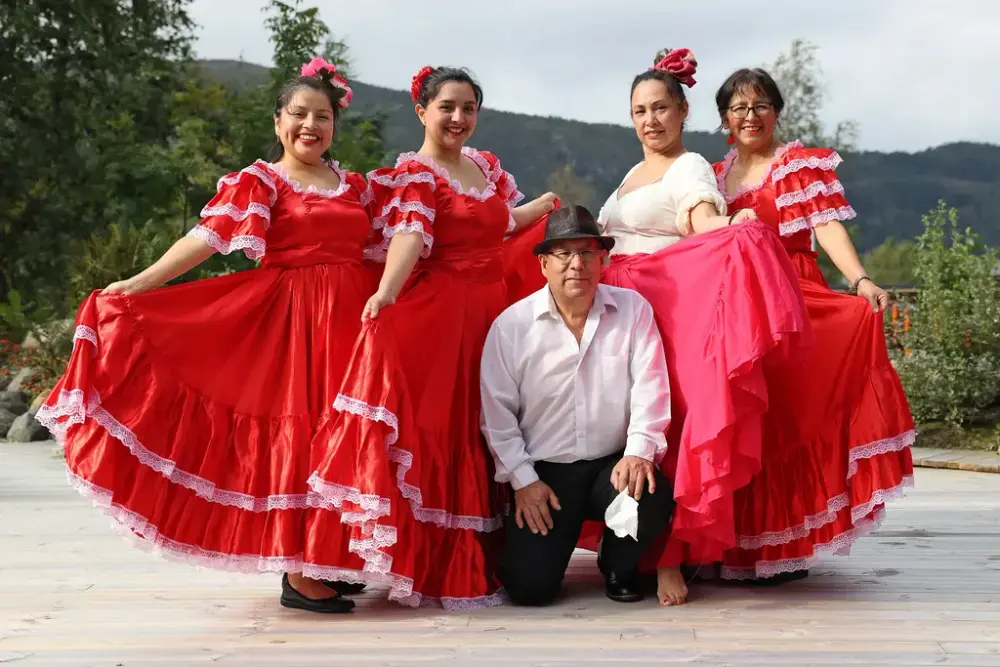 Vi ser fire damer o røde kjoler med store skjørt og en mann med hvit skjorte og hatt i midten, alle medlemmer i den peruanske folkedansgruppen Taikiri