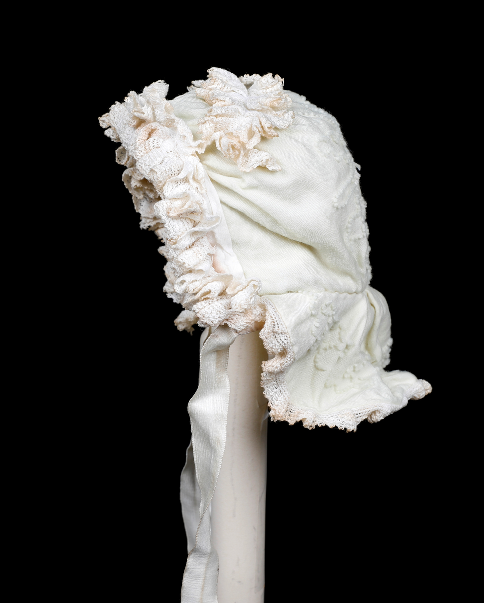 Kråke i hvit, tynn ull med ullbroderier på både pull og nakkestykke. Broderiet består av små løkker i bølgende mønster. Innfelt silkestykke på bakhodet og kanting av blonde, sannsynligvis av bomull. Luen er fôret med fint bomullslerret. Knytebånd i sateng, muligens silke.