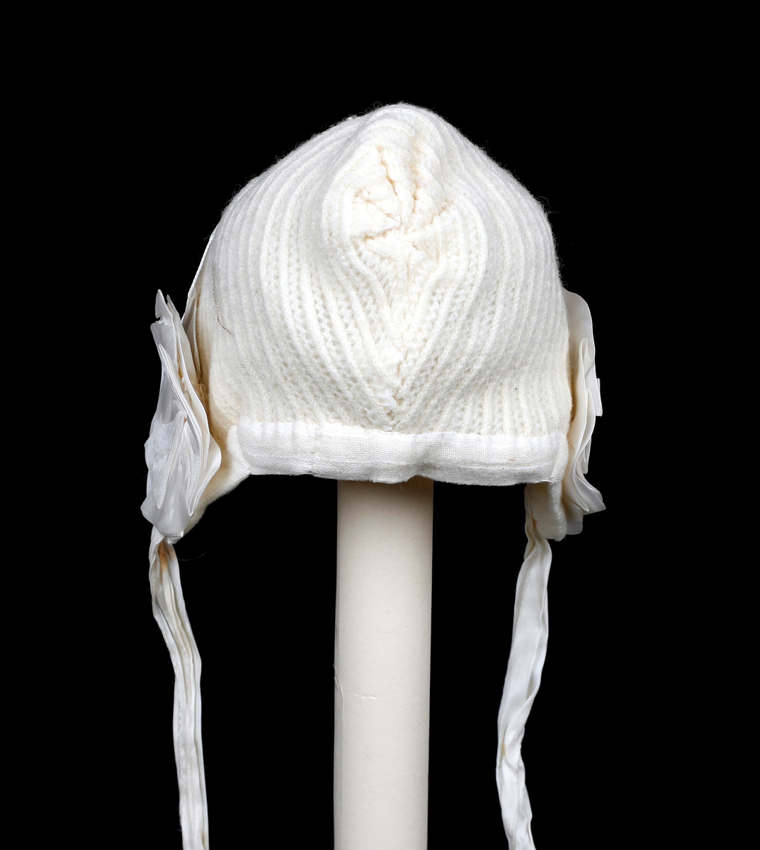Strikket lue med skru av foldet silke, silkebånd og rosetter over ørene. Fôret med gauze. Kantet i nedkant med gauze. Knytebånd i silke.