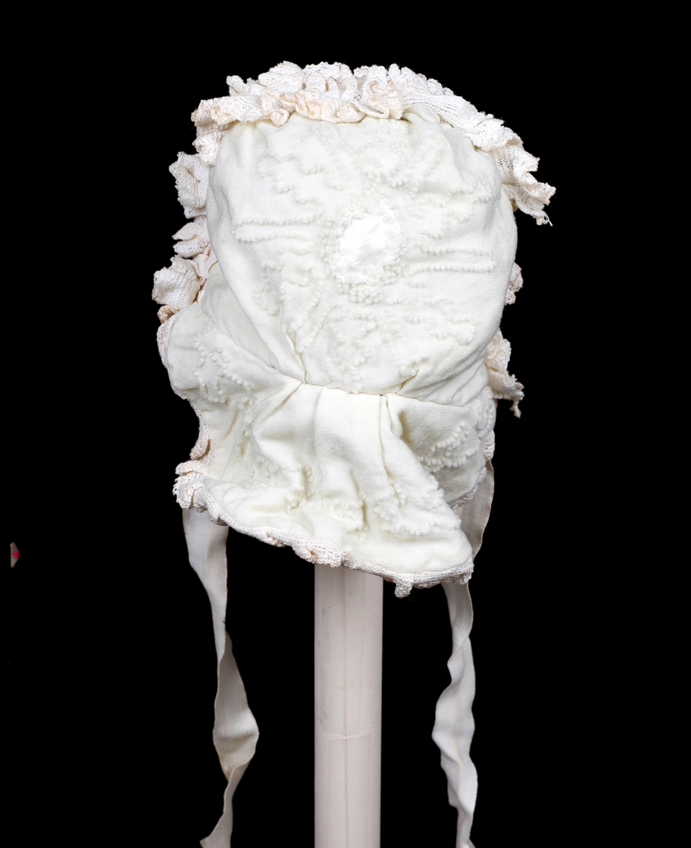 Kråke i hvit, tynn ull med ullbroderier på både pull og nakkestykke. Broderiet består av små løkker i bølgende mønster. Innfelt silkestykke på bakhodet og kanting av blonde, sannsynligvis av bomull. Luen er fôret med fint bomullslerret. Knytebånd i sateng, muligens silke.