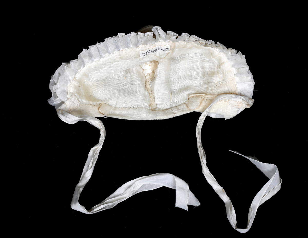 Strikket lue med skru av foldet silke, silkebånd og rosetter over ørene. Fôret med gauze. Kantet i nedkant med gauze. Knytebånd i silke.