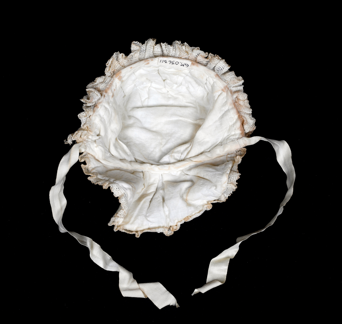 Kråke i hvit, tynn ull med ullbroderier på både pull og nakkestykke. Broderiet består av små løkker i bølgende mønster. Innfelt silkestykke på bakhodet og kanting av blonde, sannsynligvis av bomull. Luen er fôret med fint bomullslerret. Knytebånd i sateng, muligens silke.