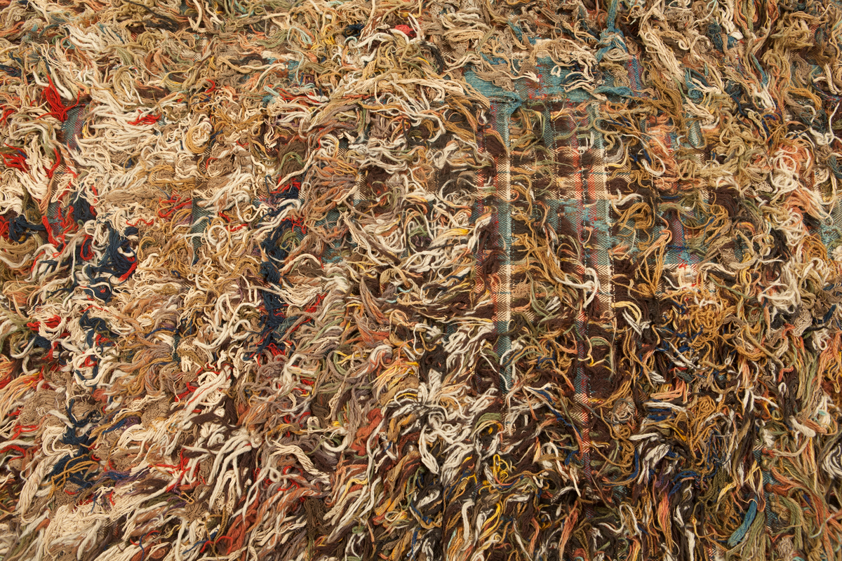 Trasrya vävd i två våder, hopsydda mot nocksidan. Rutig botten i kypert med varp av tretrådigt s-tvinnat bomullsgarn 13 tr/cm, blått med ränder i gult och brunt. Inslag av s-spunnet ullgarn, 11 inslag/cm, mörkbrunt med ränder i rött, blått och vitt. Nockor i alla färger, nockor av ullgarn, bomullsgarn och trasor. Långa med glest mellan raderna, knutna över 5 varptrådar med två varptrådar mellan nockorna och 34 inslag mellan nockraderna, 16 nockor/10 cm. 3 nockrader/10 cm.