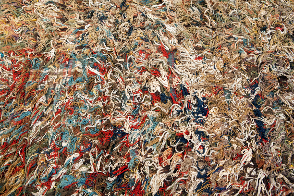 Trasrya vävd i två våder, hopsydda mot nocksidan. Rutig botten i kypert med varp av tretrådigt s-tvinnat bomullsgarn 13 tr/cm, blått med ränder i gult och brunt. Inslag av s-spunnet ullgarn, 11 inslag/cm, mörkbrunt med ränder i rött, blått och vitt. Nockor i alla färger, nockor av ullgarn, bomullsgarn och trasor. Långa med glest mellan raderna, knutna över 5 varptrådar med två varptrådar mellan nockorna och 34 inslag mellan nockraderna, 16 nockor/10 cm. 3 nockrader/10 cm.