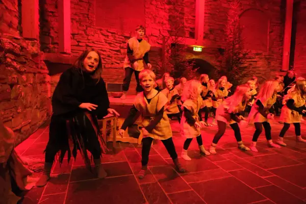 En stor flokk barn kledd i striesekker står sammen med svartkledde Lussi og speider ut over salen, helt rødt lys.