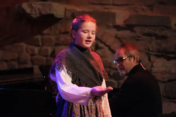 Jente med sjal og hverdagsklær fra 1800-tallet står og synger mens hun ser ned på hånda si som hun strekker fram.