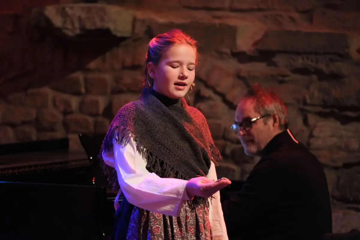 Jente med sjal og hverdagsklær fra 1800-tallet står og synger mens hun ser ned på hånda si som hun strekker fram.