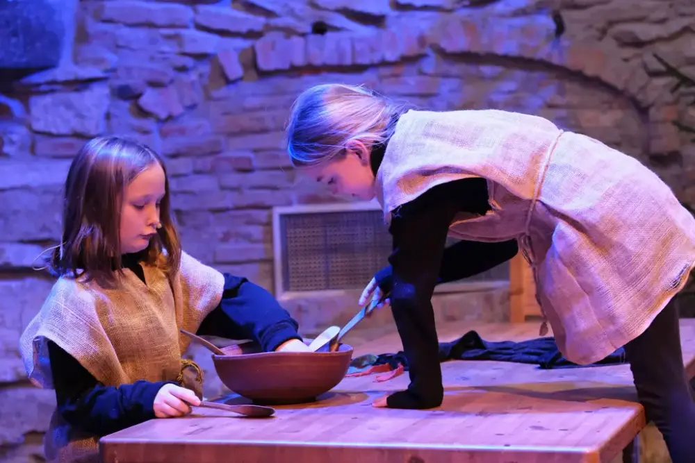 Ei jente med striesekk som klær sitter og spiser fra et stort grøtfat. Ei anna jente med striesekkjole henger inn over bordet fra andre sida ogs piser av samme grøtfatet.