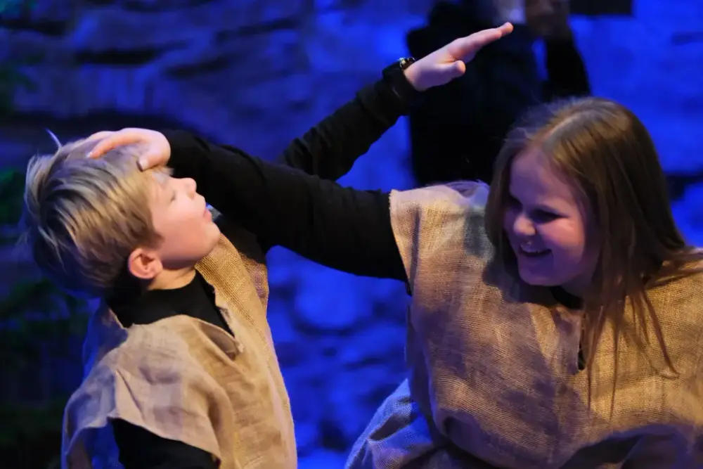 En gutt og ei jente med svarte gensere og striesekkvester utenpå holder en hånd på hverandres hode.